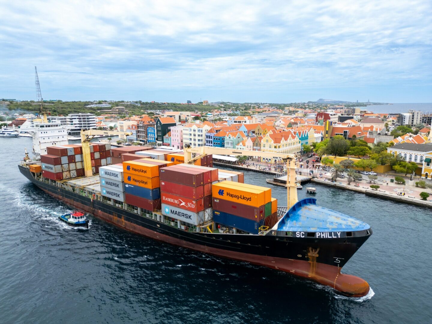 A cargo ship loaded with colorful containers sailing near a coastal town.