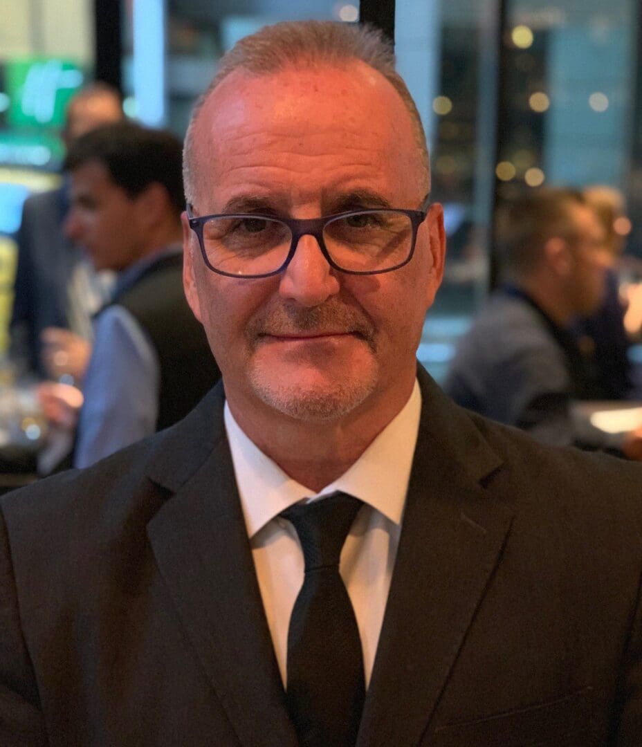 Man in glasses and suit smiling at an indoor event.