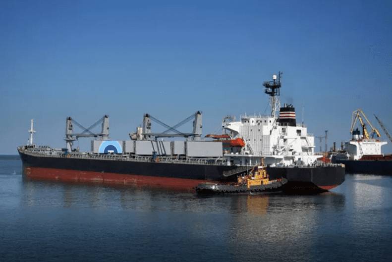 A large cargo ship docked with a tugboat beside it under clear blue sky.