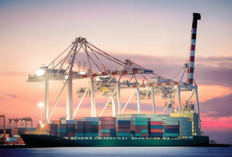 Cargo cranes unloading containers at a busy port during sunset.
